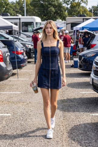A woman walking in a parking lot wearing a dark blue denim-like mini dress with a front zipper, white sneakers, and holding a coffee cup.