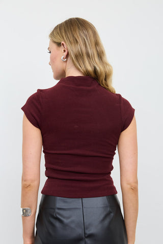A woman shown from the back wearing a fitted short-sleeve mock neck top in deep burgundy, paired with black leather-look pants, against a plain light gray studio background.