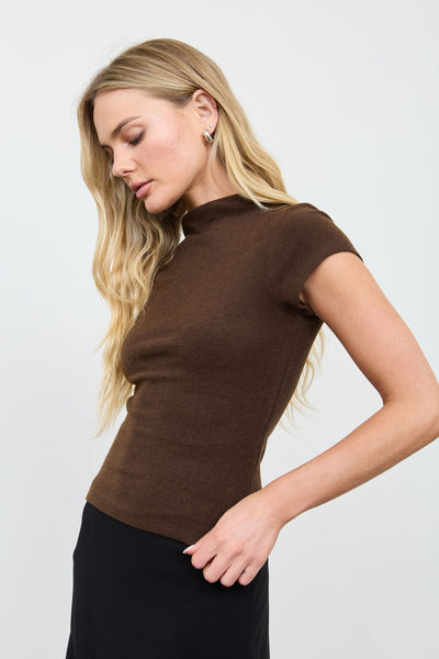 A blonde model wearing a fitted short-sleeve brown mock neck top and a black skirt, photographed against a clean light gray studio background.