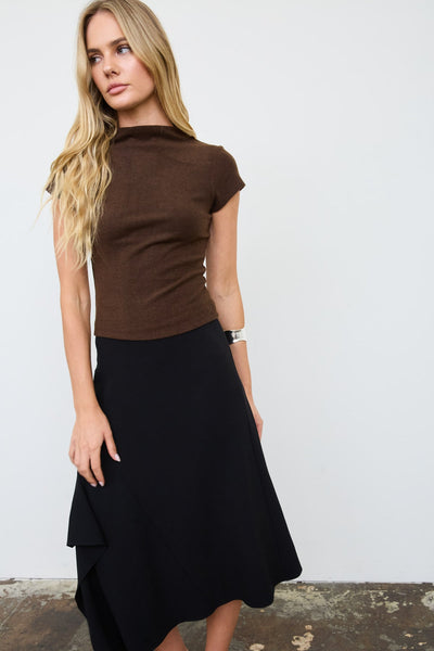 A blonde model wearing a brown short-sleeve mock neck top and a black high-low skirt, standing against a white wall.