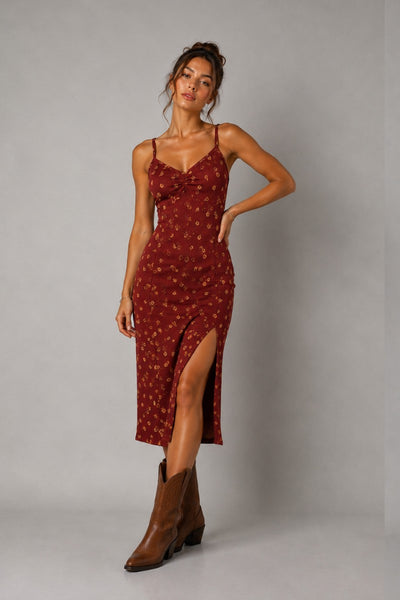 A woman models a rust-red midi slip dress with a small gold floral pattern and a thigh-high slit, paired with brown leather cowboy boots, against a neutral gray studio background.