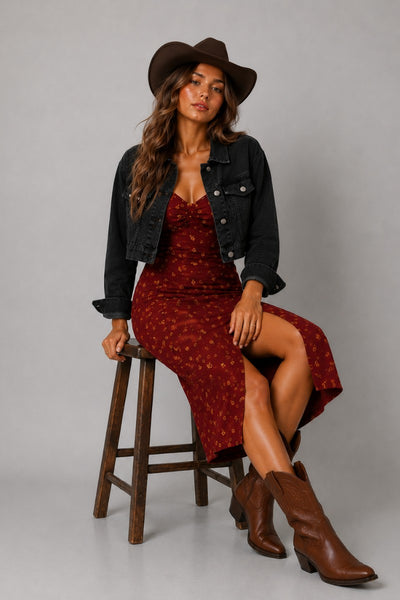A woman sits on a wooden stool wearing a dark brown cowboy hat, a black denim jacket, and a rust-red patterned midi dress with brown leather cowboy boots, photographed against a neutral gray studio background.