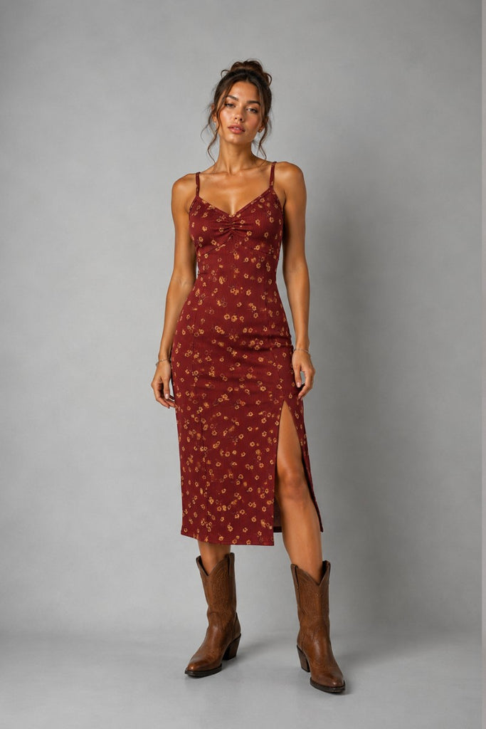 A woman modeling a rust-red spaghetti-strap midi dress with a small floral pattern and a thigh-high slit, paired with brown cowboy boots, against a solid gray studio background.