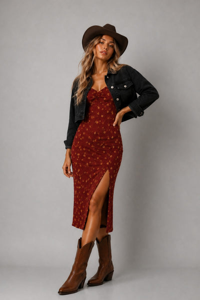 A woman modeling a red midi dress with a small yellow floral pattern, styled with a black denim jacket, a brown cowboy hat, and brown leather cowboy boots against a solid gray studio background.