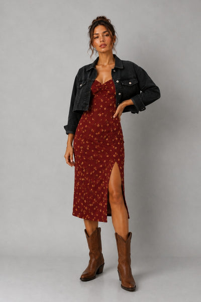 A model stands against a light gray studio background wearing a rust-red midi dress with a small floral pattern, a black denim jacket, and brown cowboy boots.