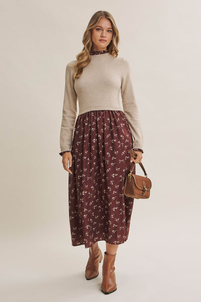 A woman modeling a beige mock-neck knit sweater paired with a dark red midi skirt featuring a small white floral pattern, holding a brown leather handbag and wearing brown ankle boots against a plain light beige studio background.