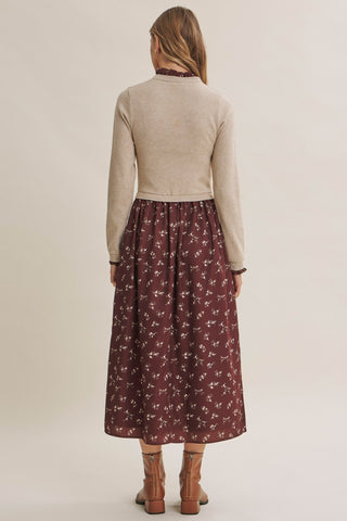 A woman shown from the back wearing a beige knit mock-neck sweater and a dark brown midi skirt with a white floral pattern, paired with brown leather boots, against a solid light beige studio background.