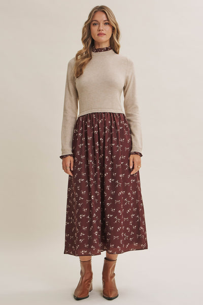 A woman modeling a beige mock-neck knit sweater paired with a dark red midi skirt featuring a small white floral print, styled with brown ankle boots against a plain beige studio background.