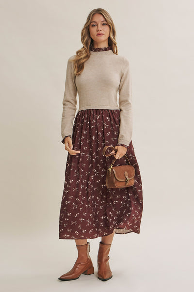 A woman models a beige mock-neck knit sweater paired with a dark brown midi skirt featuring a small white floral pattern, styled with brown ankle boots and a brown leather handbag against a solid light beige background.