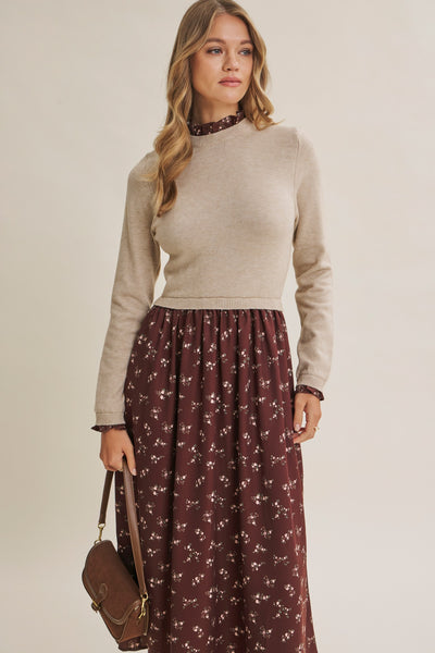 A woman modeling a beige mock-neck knit sweater paired with a dark brown floral maxi skirt, holding a brown leather handbag against a plain beige studio background.