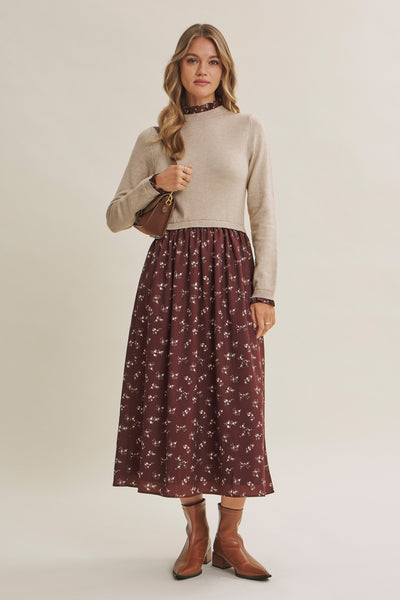A woman modeling a beige mock-neck knit sweater and a dark brown midi skirt with a small white floral print, paired with brown ankle boots and a small brown shoulder bag, against a solid light beige studio background.