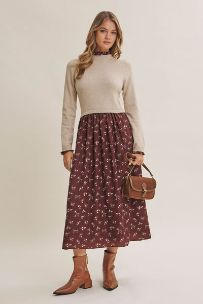 A woman modeling a beige mock-neck knit sweater paired with a dark red midi skirt featuring a small white floral pattern, holding a brown leather handbag and wearing brown ankle boots against a light beige studio background.