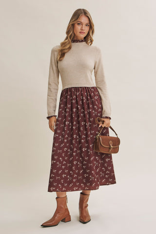 A woman modeling a beige mock-neck knit sweater paired with a dark red midi skirt featuring a small white floral pattern, holding a brown leather handbag and wearing brown ankle boots against a light beige studio background.