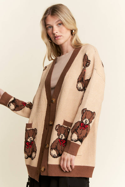 A woman wearing a beige cardigan sweater with brown trim and knitted teddy bear motifs, paired with a beige top and black pants, against a plain cream background.