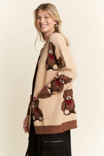 A woman wearing a beige knit cardigan with brown teddy bear patches and red bow details, paired with black pants, photographed against a plain light beige studio background.