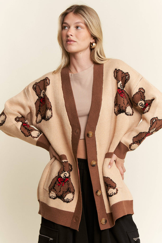 A woman wearing a beige cardigan with brown trim and knitted teddy bear motifs, styled over a beige top and black pants against a plain cream background.