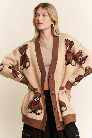 A woman wearing a beige cardigan with brown trim and knitted teddy bear motifs, styled over a beige top and black pants against a plain cream background.