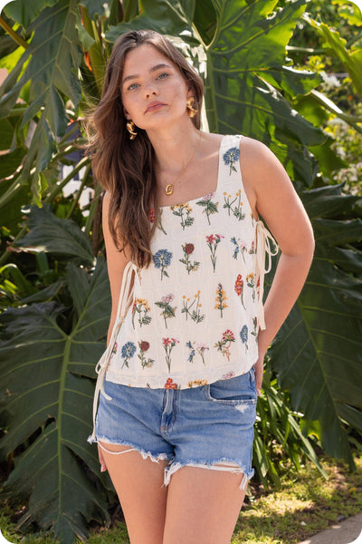 Woman wearing a floral tank top and denim shorts standing in front of large green leaves.