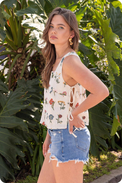 Woman wearing a floral top and denim shorts standing in front of green foliage