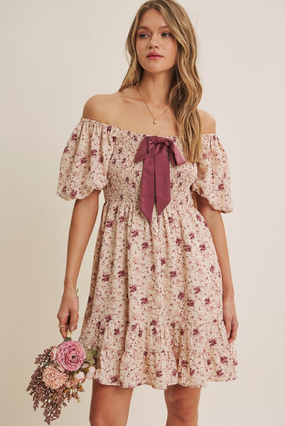 A woman modeling an off-the-shoulder floral mini dress with a dusty rose bow at the neckline, holding a small bouquet of pink and purple flowers against a plain beige background.