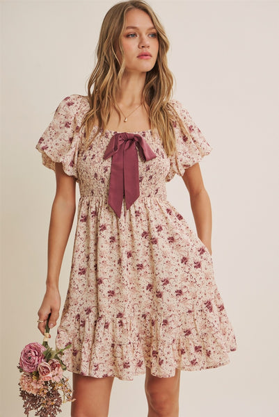 A woman modeling a short, knee-length floral dress with puff sleeves, a square neckline, and a large dusty rose bow at the center front. She is holding a small bouquet of pink flowers against a plain beige studio background.