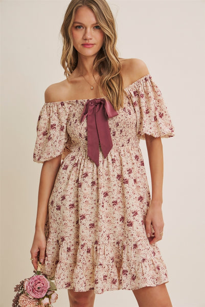 A woman models an off-the-shoulder floral mini dress with a dusty rose ribbon bow at the neckline, set against a plain beige background.