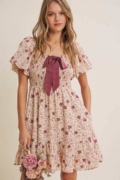 A woman wearing a short-sleeve floral dress with a large dusty rose bow at the neckline, holding a small bouquet of pink flowers against a plain beige studio background.