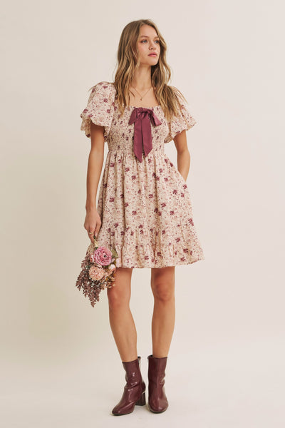 A woman modeling a short floral dress with flutter sleeves and a dark mauve bow at the neckline, holding a small bouquet of flowers, against a plain light beige studio background.