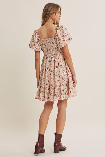 A woman modeling a short floral dress from the back, featuring puff sleeves, a square neckline, and a flared skirt. She is wearing dark brown heeled ankle boots against a plain beige studio background.