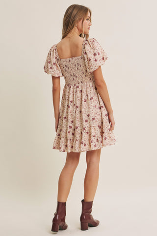 A woman modeling a short floral dress from the back, featuring puff sleeves, a square neckline, and a flared skirt. She is wearing dark brown heeled ankle boots against a plain beige studio background.