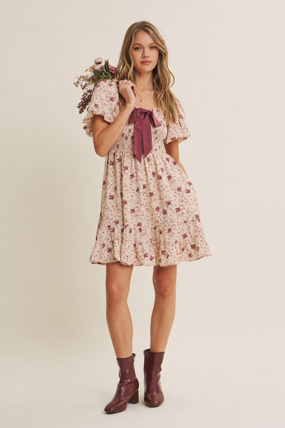 A woman modeling a short puff-sleeve floral dress with a dark mauve bow at the neckline, holding a bouquet of flowers, set against a plain beige studio background.