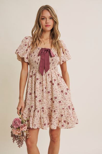 A woman modeling a short puff-sleeve dress with a small floral print in pink and burgundy on a cream base, featuring a large burgundy bow at the neckline. She is holding a small bouquet of pink flowers against a plain beige studio background.