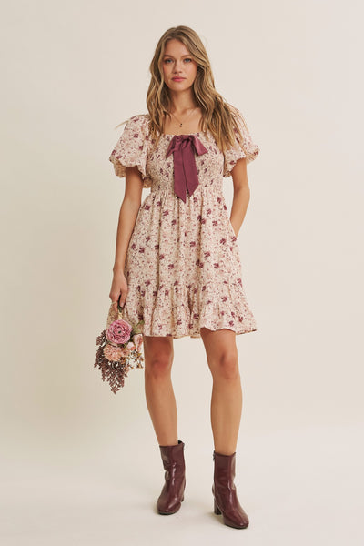 A woman modeling a short puff-sleeve floral dress with a deep burgundy bow at the neckline, holding a small bouquet of pink and purple flowers. She is wearing burgundy ankle boots against a plain beige studio background.