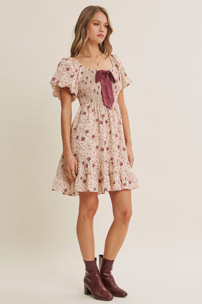 A woman modeling a short floral dress with puff sleeves, a square neckline, and a dark mauve bow at the center front, paired with dark mauve ankle boots, against a plain beige studio background.