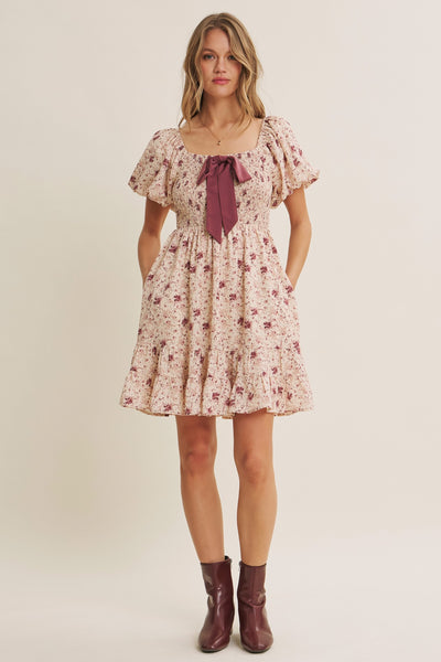 A woman modeling a short puff-sleeve floral dress with a deep burgundy bow at the neckline, paired with burgundy ankle boots, against a plain beige studio background.