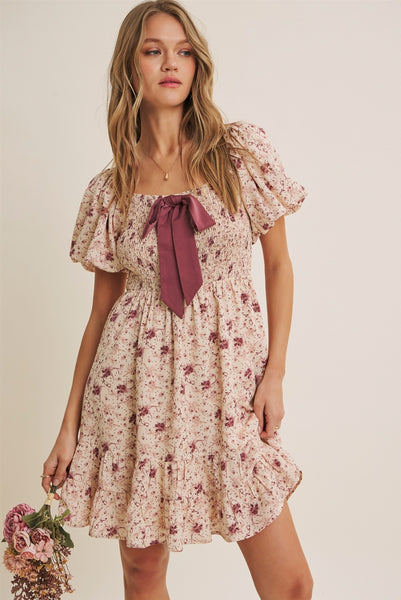 A woman modeling a short puff-sleeve floral dress with a dusty rose bow at the neckline, set against a plain beige background.