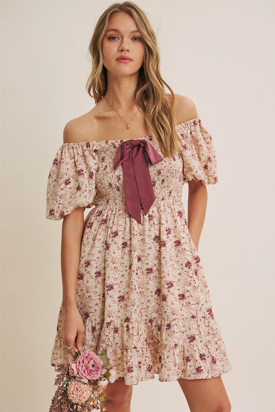 A woman modeling an off-the-shoulder floral dress with a dusty rose bow at the neckline, set against a plain beige background.