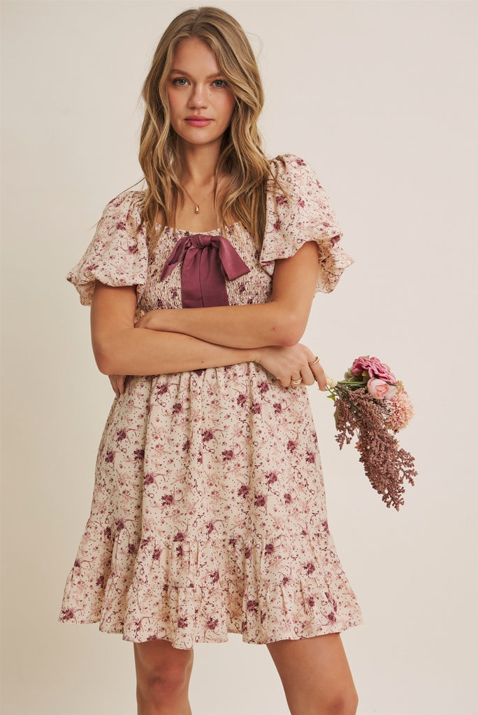 A woman modeling a short-sleeve floral dress with a ruffled bodice, square neckline, and a dark mauve bow at the center chest. She is holding a small bouquet of pink and purple flowers against a plain beige studio background.