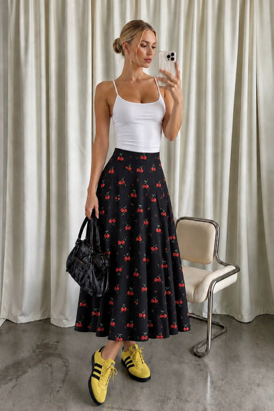 A woman takes a mirror selfie while wearing a black midi skirt with a red cherry print, a white spaghetti-strap tank top, and yellow sneakers. She is holding a black handbag, standing in front of a beige curtain with a vintage chair to her side.