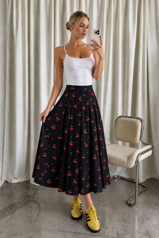 A woman takes a mirror selfie wearing a white ribbed tank top and a black midi skirt with a red cherry print. She is standing in front of beige curtains, with a vintage-style chair to her right.