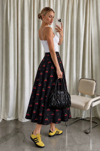 A woman takes a mirror selfie wearing a white ribbed tank top and a black midi skirt with a red cherry print. She is holding a black quilted handbag and wearing yellow sneakers, standing in front of beige curtains.