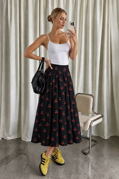 A woman takes a mirror selfie wearing a white ribbed tank top and a black midi skirt with a red cherry print. She is holding a black handbag and wearing yellow sneakers, standing in front of a beige curtain with a modern chair to the side.