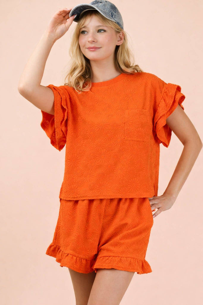 Woman wearing an orange outfit with ruffled sleeves and shorts on a beige background