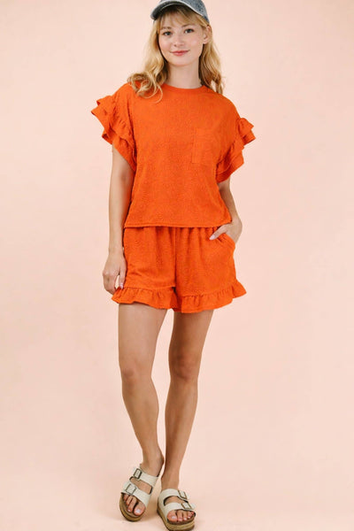 Young girl wearing an orange outfit with ruffled sleeves and shorts on a pink background