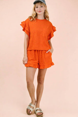 Young girl wearing an orange outfit with ruffled sleeves and shorts on a pink background