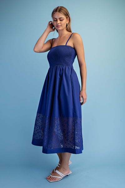 A woman modeling a royal blue midi dress with thin straps, a smocked bodice, and a lace-trimmed hem, standing against a solid light blue studio background.