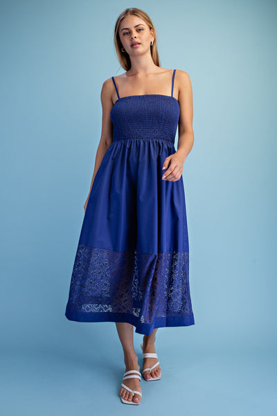 A woman modeling a blue midi dress with thin straps, a smocked bodice, and a lace-trimmed hem, standing against a solid light blue studio background.