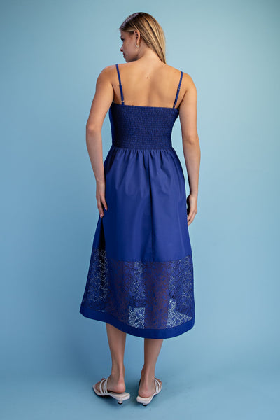 A woman viewed from the back wearing a royal blue midi dress with thin straps, a smocked bodice, and a lace-trimmed hem, standing against a solid light blue studio background.