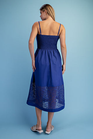 A woman viewed from the back wearing a royal blue midi dress with thin straps, a smocked bodice, and a lace-trimmed hem, standing against a solid light blue studio background.