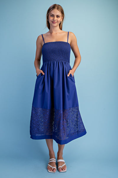 A woman modeling a blue midi dress with thin straps, a smocked bodice, side pockets, and a lace hem, standing against a solid light blue background.